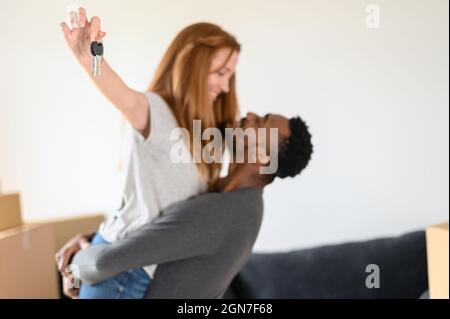 Fuoco selettivo alle chiavi della porta in mano della donna. Felice coppia mista-corsa che si sposta in nuova casa, l'acquisto del proprio bene immobile, si guarda a vicenda eccitato, coniugi internazionali in movimento Foto Stock