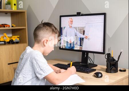Il ragazzo studia di fronte al computer, partecipando a un corso di formazione che conduce un insegnante senior, prende appunti. E-learning, studio on-line a casa, scuola a casa Foto Stock