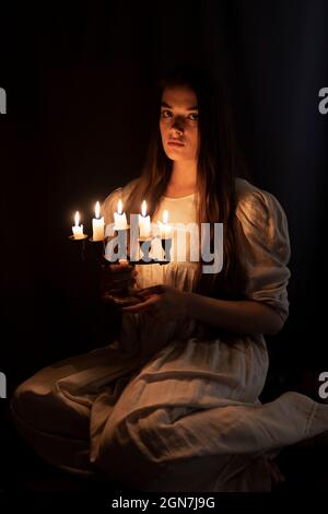 Una giovane ragazza in un vecchio vestito bianco seduto e tenendo una candela nelle sue mani. Sfondo scuro. Spaventoso concetto di orrore. Foto Stock