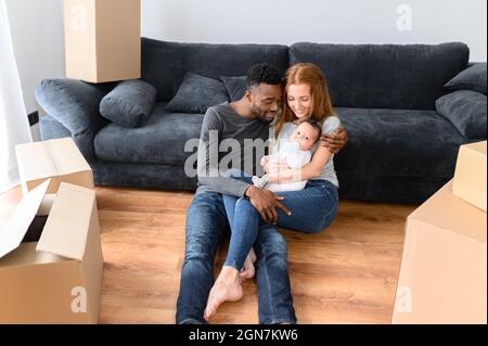 Felice famiglia internazionale misto-corsa durante la giornata di disimballaggio, in movimento nella nuova casa. Il padre africano, la madre caucasica e la figlia del bambino biraciale carina che giacciono sul pavimento tra le scatole di cartone Foto Stock
