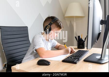 Il ragazzo che indossa le cuffie sta studiando davanti al computer, prende appunti, ascolta musica e scrivi con una penna, preparandosi all'esame. E-learning, studio a casa online, homeschooling Foto Stock