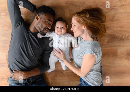Happy misto-corsa internazionale famiglia di tre che si trovano sul pavimento, felice marito nero, moglie caucasica e la loro bambina biraciale guardando la macchina fotografica, vista dall'alto Foto Stock