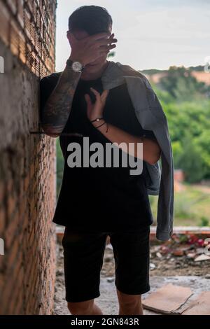 Ritratto di giovane bel caucasico in abiti casual neri, coprendo il volto con mano. Giacca in denim, tatuaggio completo con braccio Foto Stock