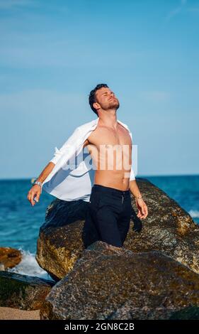 Uomo sereno in elegante camicia bianca e con corpo in piedi con occhi chiusi sulla costa rocciosa e godere della libertà nelle giornate di sole Foto Stock