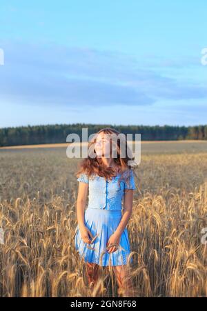 La ragazza giovane felice sta sorridendo al campo di grano, toccando le orecchie di grano con la sua mano. Bellissimo adolescente godendo la natura in caldo sole in un campo di grano Foto Stock