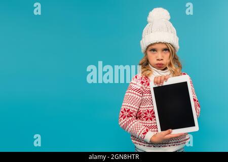 annoiato bambino in caldo cappello e maglione che tiene tablet digitale con schermo vuoto isolato su blu Foto Stock