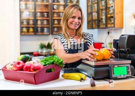 Donna di vendita sorridente che pesa arance sulla bilancia al banco del check-out del negozio Foto Stock