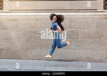 Donna felice con zaino che salta sul sentiero di fronte al muro Foto Stock