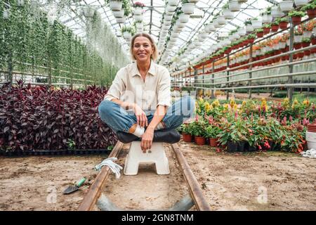 Donna contadina sorridente seduta a gambe incrociate su sgabello in serra Foto Stock
