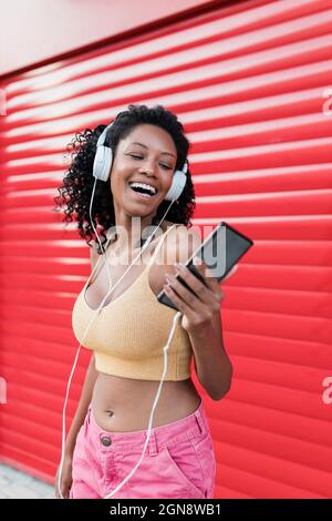 Afrodonna che ascolta la musica con le cuffie mentre ride con l'otturatore rosso Foto Stock