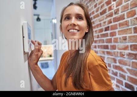 Sorridente donna che contempla l'utilizzo di un dispositivo di automazione domestica a parete Foto Stock