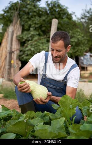 Coltivatore maschio sorridente che tiene squash mentre accovacciato a campo agricolo Foto Stock