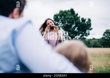 Donna sorridente che fotografa la famiglia attraverso la macchina fotografica al parco Foto Stock