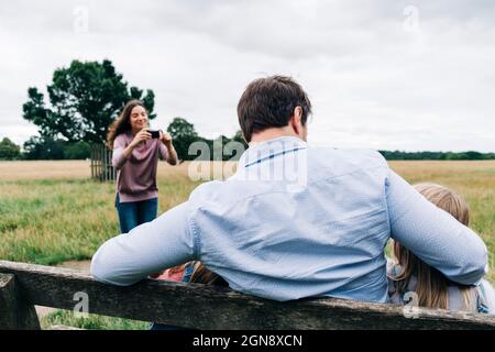 Donna che fotografa l'uomo seduto con le figlie su panca attraverso la macchina fotografica Foto Stock