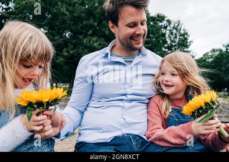 Figlie che tengono girasoli mentre si siedono con il padre al parco Foto Stock