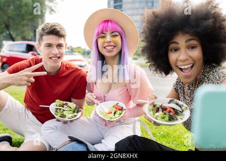 Donna allegra che prende selfie con gli amici mentre l'uomo mostra segno di pace Foto Stock