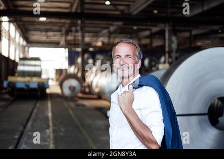 Un uomo d'affari sorridente che tiene un blazer mentre si trova nell'industria del metallo Foto Stock