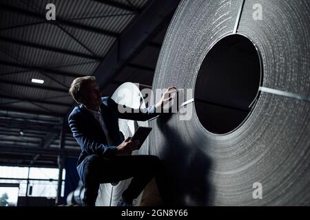 Ispettore maschio che controlla la lamiera d'acciaio arrotolata durante l'accovacciamento in magazzino Foto Stock