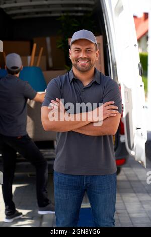 Uomo di consegna sorridente con le braccia incrociate in piedi vicino a un furgone in movimento Foto Stock