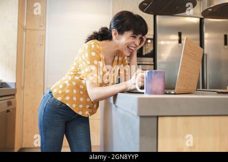 Donna matura ridendo mentre videochiama attraverso il laptop durante gli incontri online Foto Stock