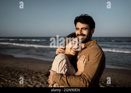 Felice coppia giovane abbracciarsi mentre si sta in piedi in spiaggia Foto Stock