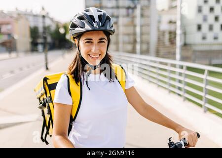 Donna di servizio essenziale sorridente che indossa casco da ciclismo e zaino di consegna il giorno di sole Foto Stock