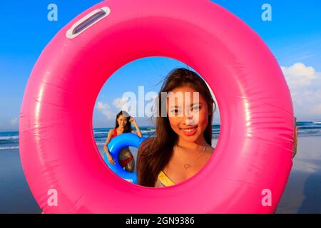 Sorridente giovane donna che guarda attraverso anello gonfiabile con un'amica in background alla spiaggia Foto Stock