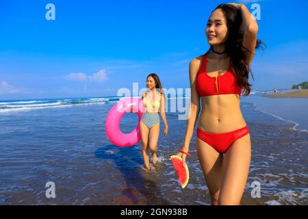 Donna sorridente che tiene cocomero mentre amico femmina con galleggiante di nuoto che cammina attraverso l'acqua in giorno di sole Foto Stock