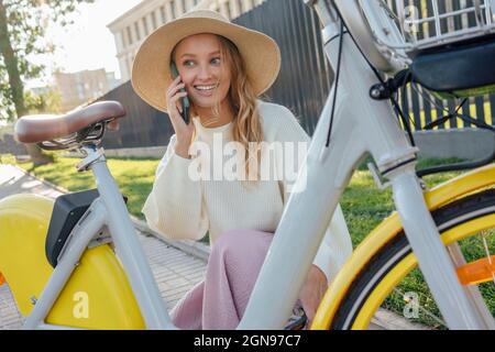 Donna bionda sorridente che parla sullo smartphone mentre si accovacciano in bicicletta Foto Stock