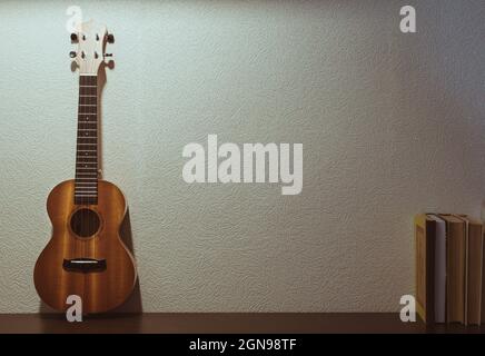 Un ukulele in legno si erge diritto contro il muro e vecchi libri, spazio copia Foto Stock