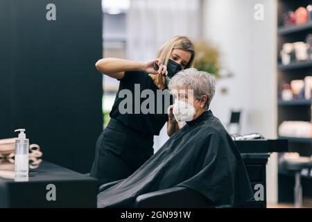 Donna parrucchiere taglio capelli del cliente in salone durante COVID-19 Foto Stock