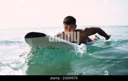 Il giovane uomo che si addita in acqua su una tavola da surf in una giornata di sole. Foto Stock