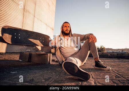 ragazzo in tuta grigia con cappuccio seduto sul pavimento appoggiato su uno skateboard al tramonto Foto Stock