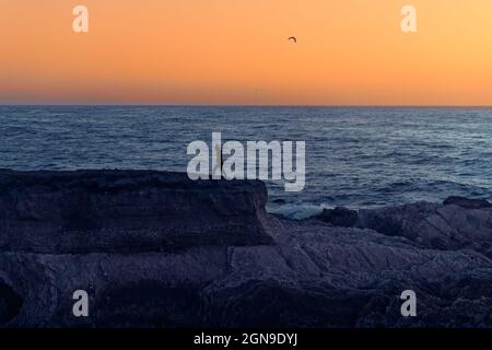 Mare al tramonto. Scogliere rocciose e silhouette di donna camminata, oceano blu e cielo giallo brillante sullo sfondo, spazio copia Foto Stock