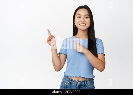 Sorridente ragazza asiatica che punta a sinistra e mostra banner di vendita, annunciando l'offerta promozionale, in piedi in maglietta su sfondo bianco Foto Stock