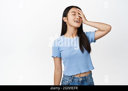 Bellezza e cura della pelle. Spensierate donna asiatica che tocca il viso, gli occhi stretti e sorridente sognante, in piedi in t-shirt blu su sfondo bianco Foto Stock