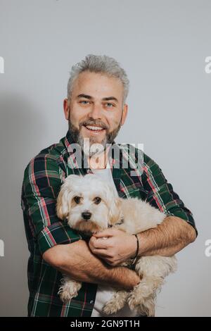 Uomo multirazziale adulto con capelli bianchi che tiene un adorabile cane maltese contro un muro grigio Foto Stock