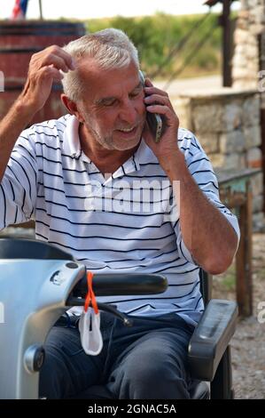 Uomo anziano seduto su sedia a rotelle elettrica e con telefono cellulare Foto Stock
