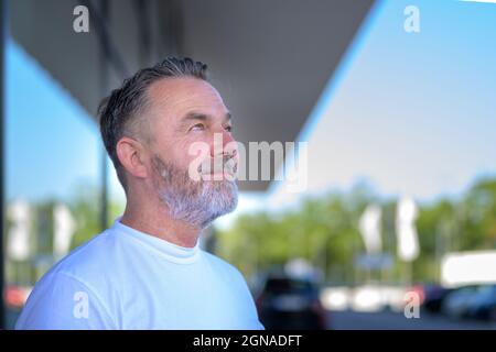Felice uomo anziano bearded che guarda in su al cielo con un sorriso di fascio di piacere e di contentment mentre si alza fuori di un edificio commerciale in estate i Foto Stock
