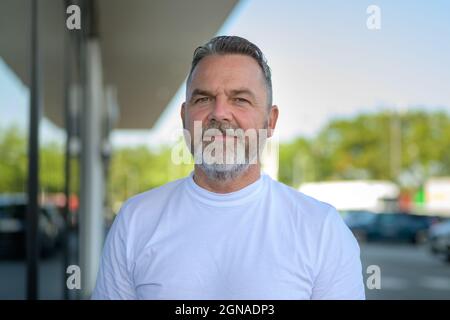Uomo anziano con bearded in piedi in un parcheggio esterno che guarda la macchina fotografica con un'espressione pensiva tranquilla in un ritratto della testa e delle spalle Foto Stock