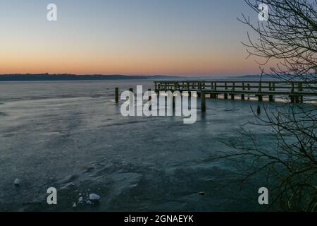 Molo di legno e mucchi in attesa di primavera in un lago ghiacciato in una giornata fredda all'alba, paesaggio tranquillo, spazio copia, fuoco selezionato Foto Stock
