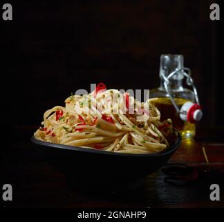 Ciotola con deliziosi spaghetti aglio e olio con erbe e peperoncino rosso servito con bottiglia di olio d'oliva su sfondo nero in studio Foto Stock