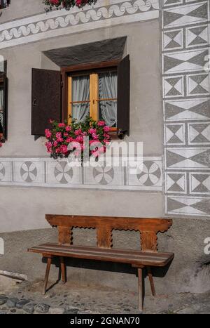 Un luogo soleggiato in cui sedersi in un angolo di un'antica casa restaurata a Scuol Sot, il centro storico di Scuol, Graubünden o Grigioni cantone, Svizzera, con una panca in legno con uno schienale intagliato in piedi su ciottoli sotto fasce di decorazione sgraffito e fiori rosa e rosso luminosi che sbocciano in un box finestra. Foto Stock