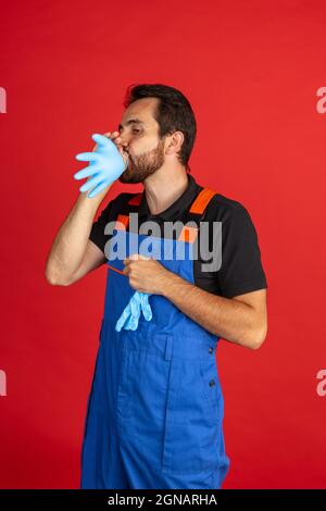 Ritratto comico di giovane uomo barbuto, meccanico maschile auto o calzatore indossando dungarees lavoro blu isolato su sfondo rosso studio. Concetto di viso Foto Stock