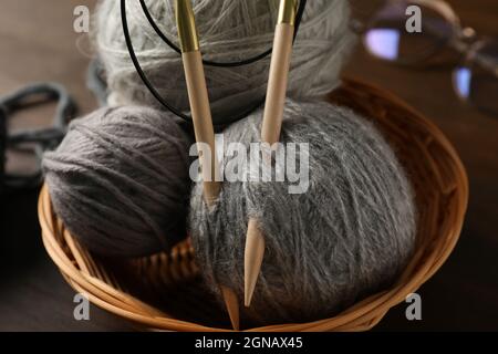 Ciotola di vimini con palle di filato e aghi da maglieria, e bicchieri su sfondo di legno Foto Stock