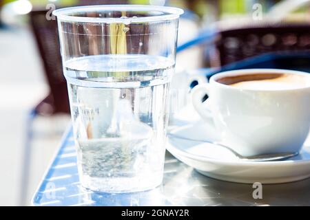 Vetro trasparente di plastica con acqua e tazza di cappuccino e tavolini di caffe sullo sfondo sfocati. Caffè servito in stile italiano tradizionale. Ancora Foto Stock