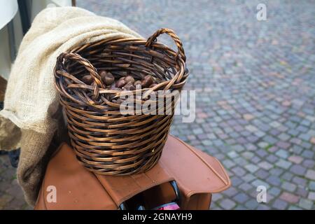 Cestino di vimini marrone con due manici pieni di castagne messo sul portabiciclette in vendita. Prodotti agricoli. Coperta di biancheria e pavimentazione di ciottoli Foto Stock