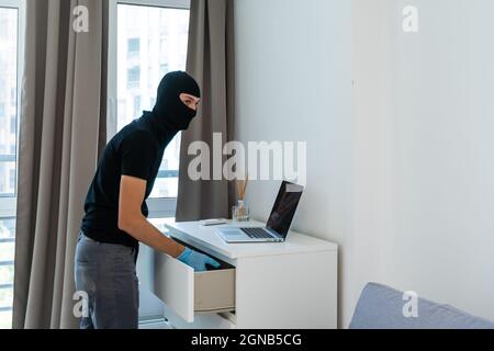 Ladro con balaclava nera rubare laptop. Il burglar commette un crimine in appartamento di lusso Foto Stock