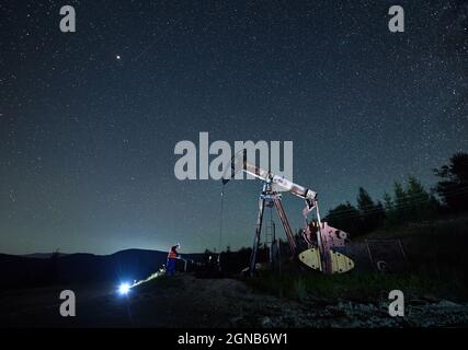 L'oilman che lavora il turno di notte nel pozzo di petrolio. Operatore che controlla i parametri di funzionamento del martinetto della pompa dell'olio. Sullo sfondo un bel cielo stellato e silhouette colline di montagna. Foto Stock