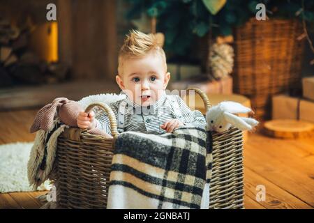 Bambino di un anno con il suo giocattolo preferito in un cestino di vimini sul pavimento vicino all'albero di Natale e regali. Stagione di festa. Vigilia di Capodanno di Chris Foto Stock